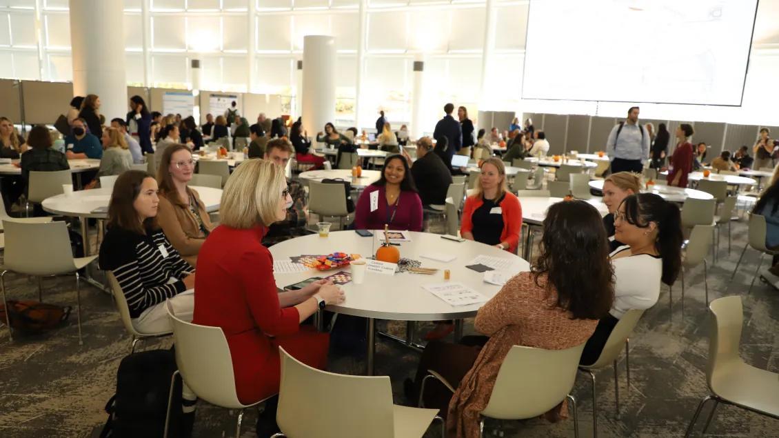 People seated around tables engaged in discussions in a large room with a large display screen in the back.