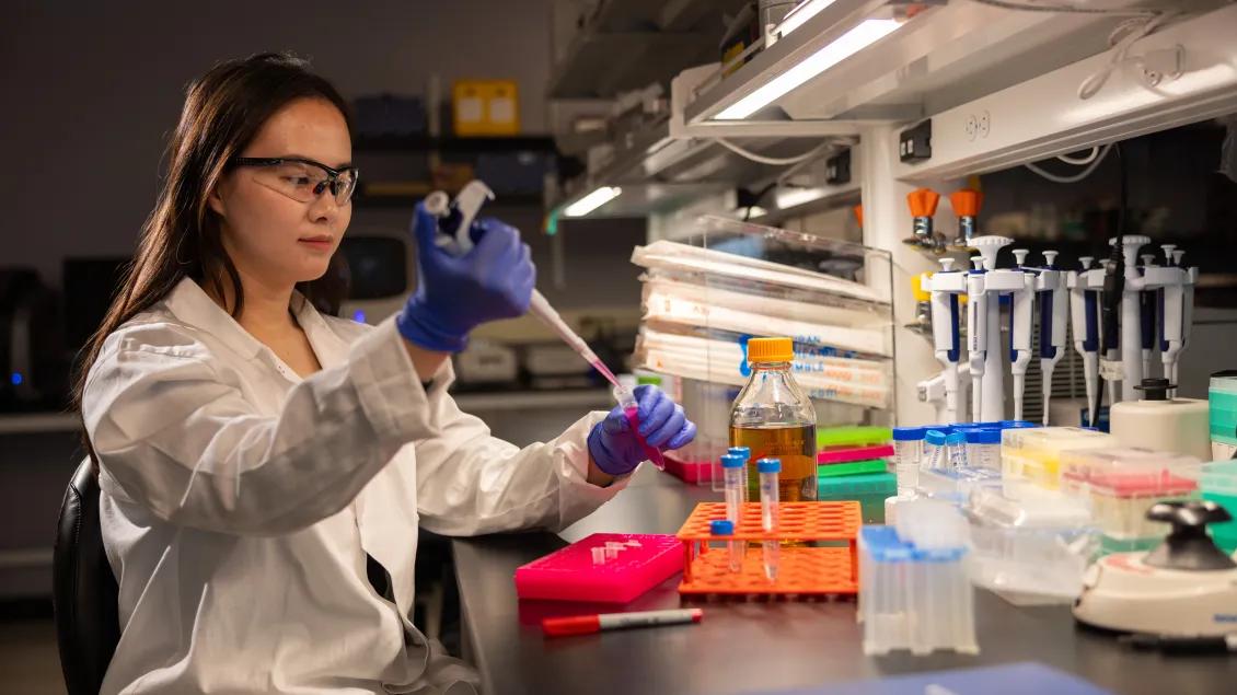 A person sits in white lab coat using a pipette while working in a research lab.