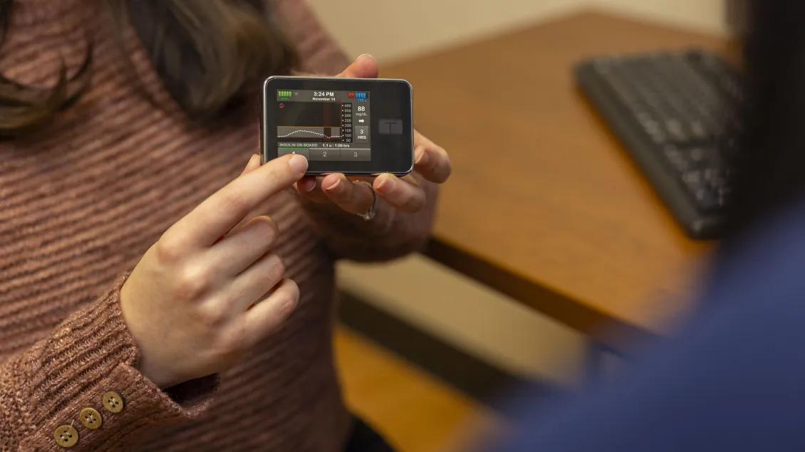 Person holding and pointing to a continuous glucose monitor receiver displaying glucose trend data
