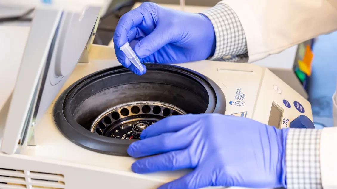A gloved hand places a small tube into a lab centrifuge.
