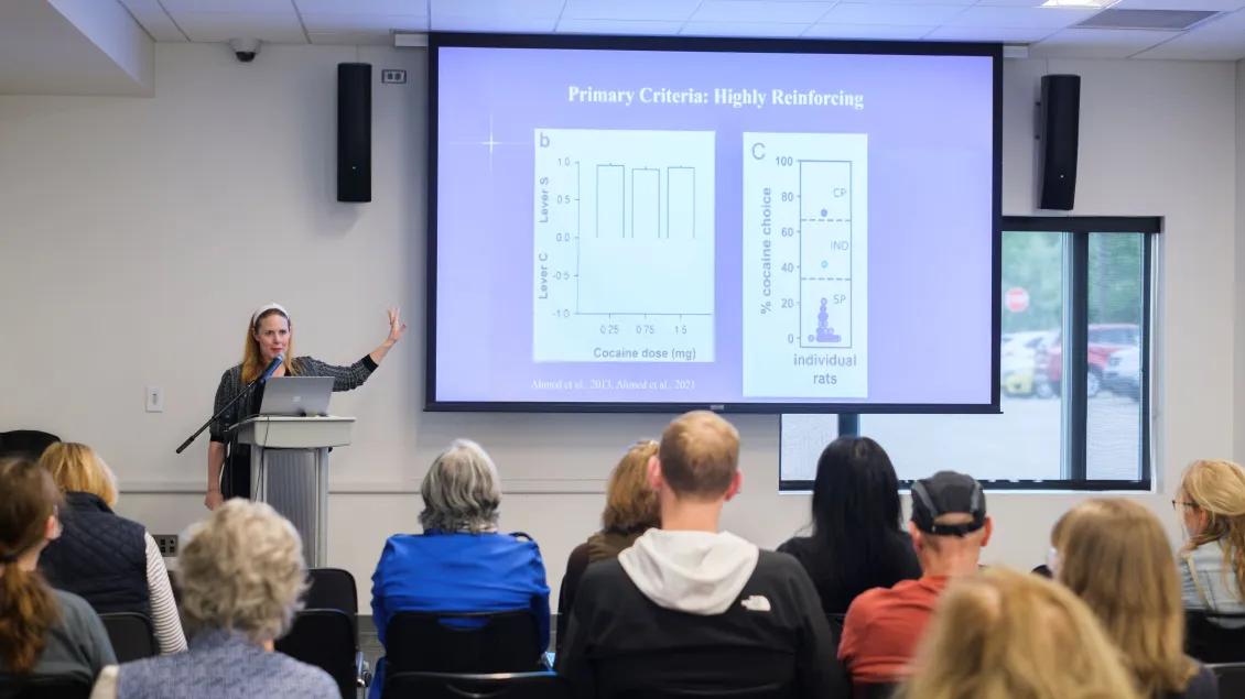 Someone standing at a podium presenting in front of an audience with a large screen to the left of them. There are 