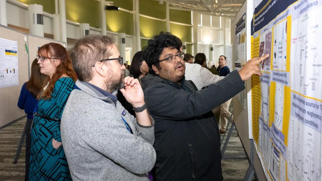 two people looking at a research poster with one pointing directly at something on the poster.