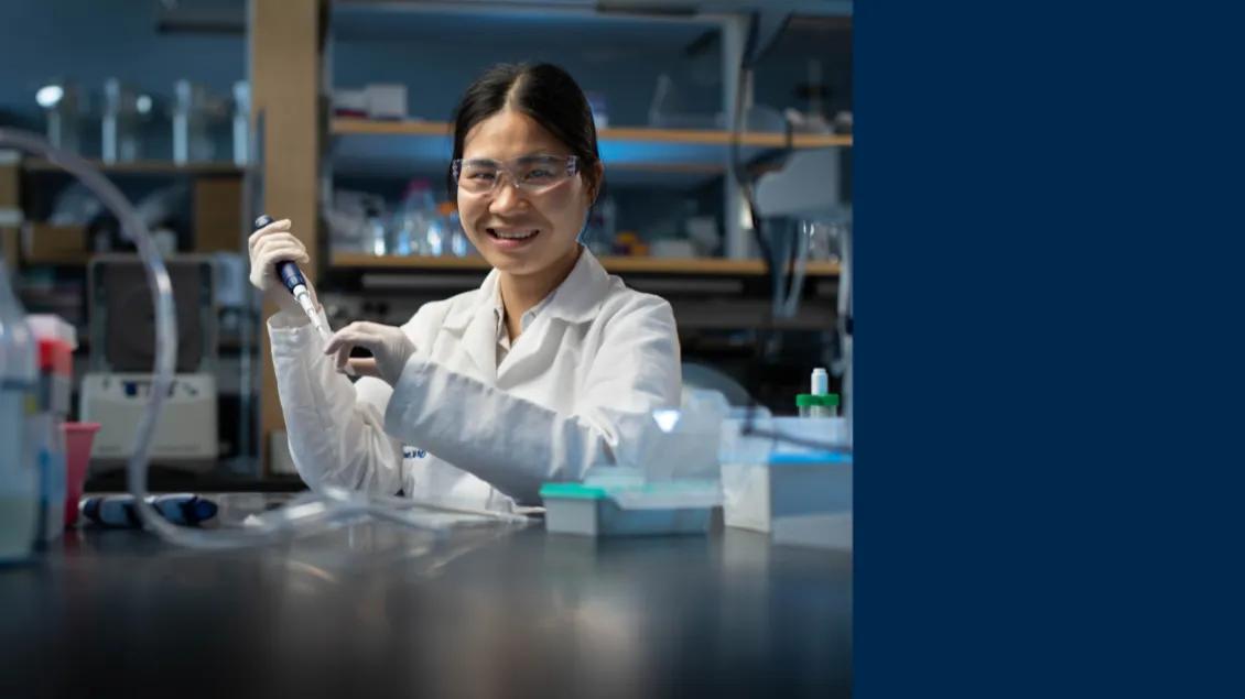 A person smiling wearing a white coat and protective glasses holds a pipette while working in a lab