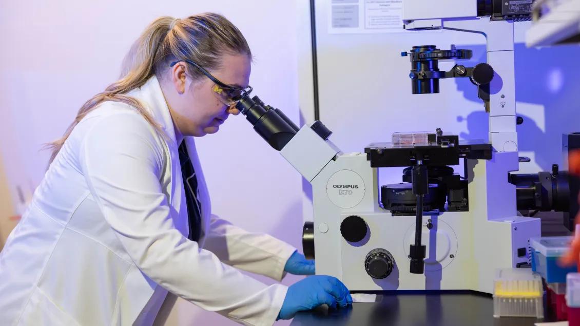 A person wearing a white coat and blue gloves looks into a microscope.