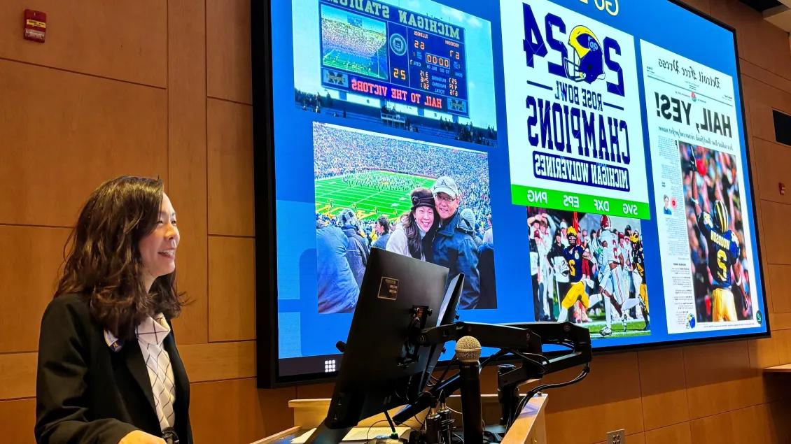 Frontiers Seminar speaker presenting at a podium in a lecture hall, with a presentation slide displayed on a large screen showing personal photos and University of Michigan football-related images.