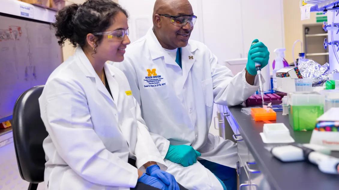 Two people in white lab coats and safety goggles pipetting. 