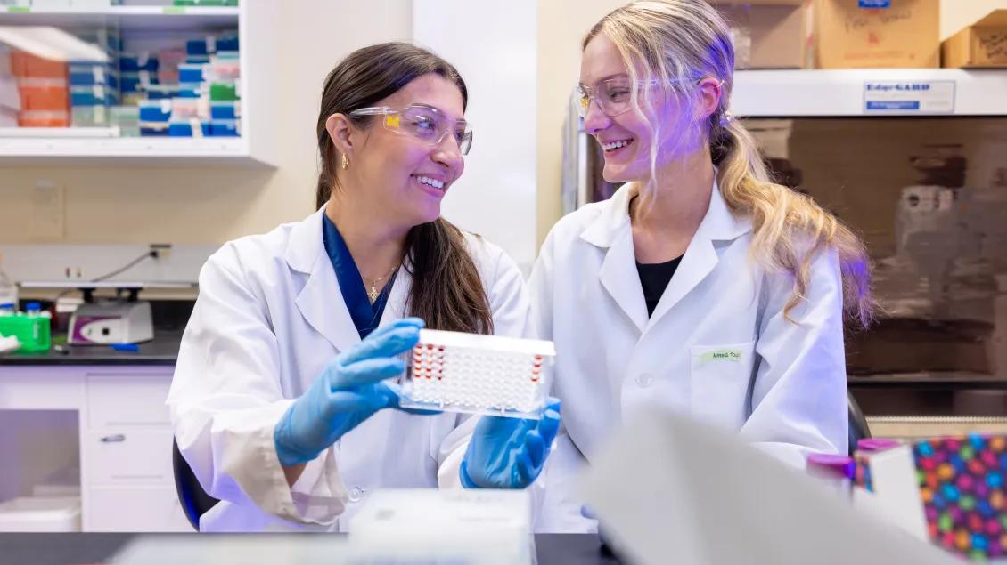 Two women wearing white lab coats and safety goggles smile while looking at a set of specimens.