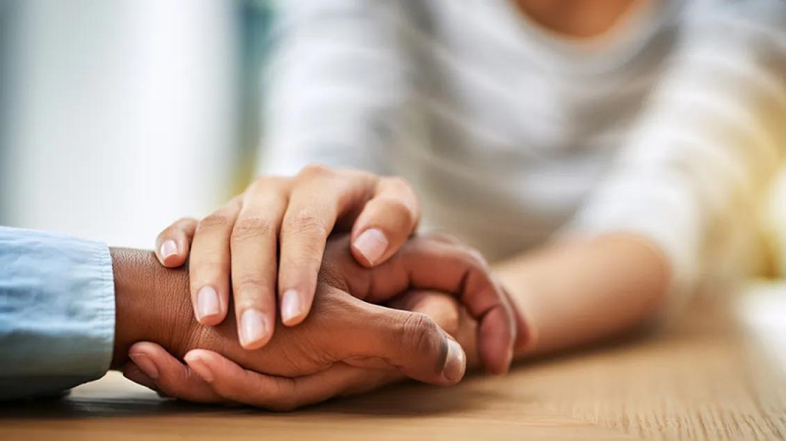 Two hands rest gently on one another on a wooden table, conveying support and compassion.
