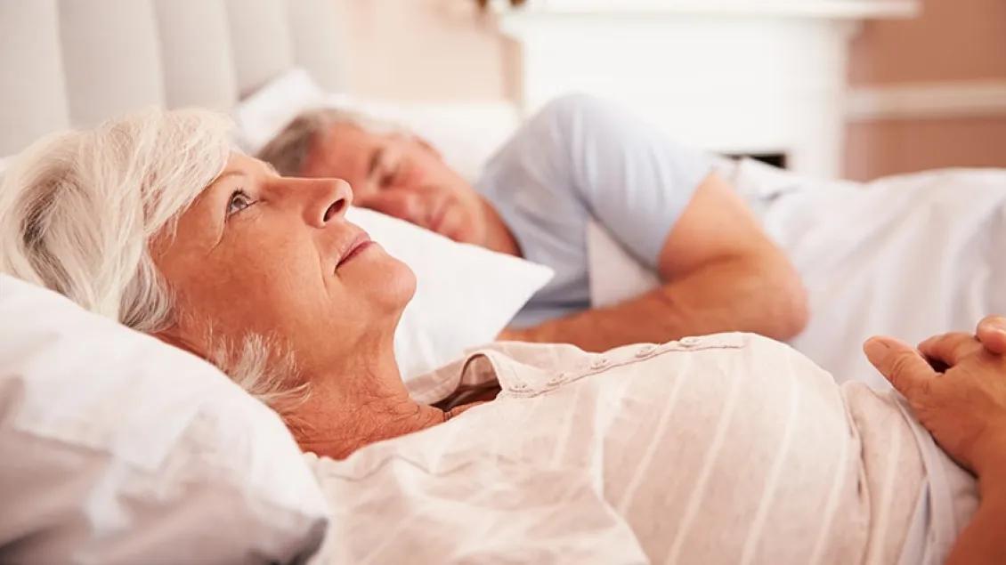 Elderly woman lying awake in bed next to a sleeping man, looking contemplative.