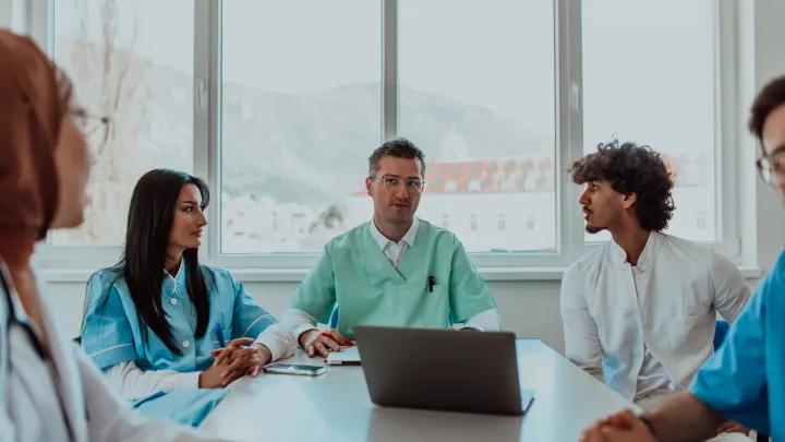 A group of multiethnic medical professionals including doctors, surgeons, and nurses are gathered in a hospital setting discussing patient care.