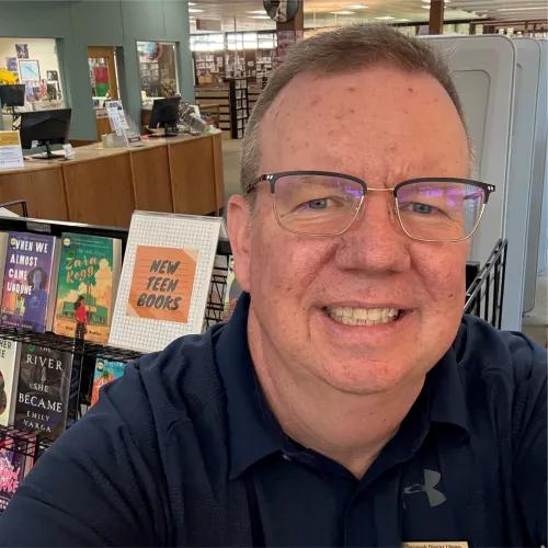 Close-up selfie of a smiling man wearing glasses and a dark polo shirt in a library, with a “New Teen Books” display and service desk behind him.