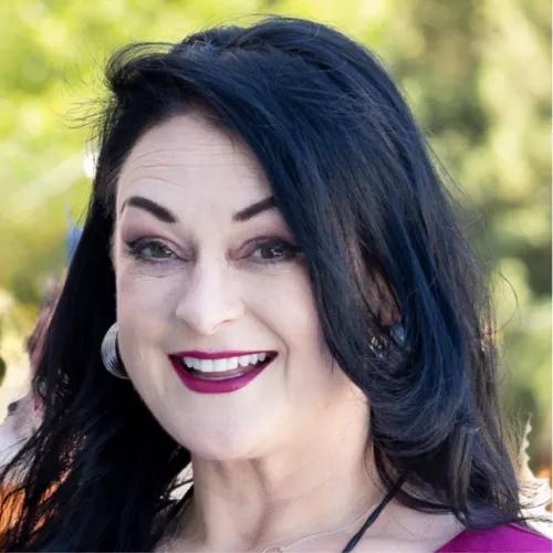  Close-up outdoor portrait of a smiling woman with long dark hair, hoop earrings, and bright pink lipstick against a softly blurred green background.
