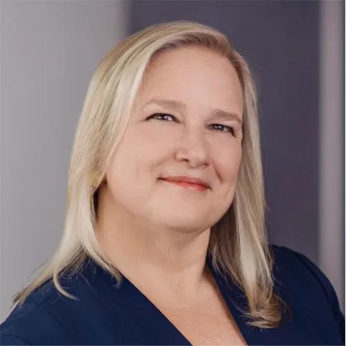 Professional headshot of a smiling woman with shoulder-length blonde hair wearing a navy blouse against a softly blurred gray background.