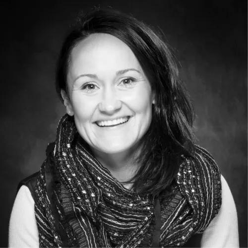 Black-and-white headshot of a smiling woman with long dark hair, wearing a textured patterned scarf against a dark studio backdrop.