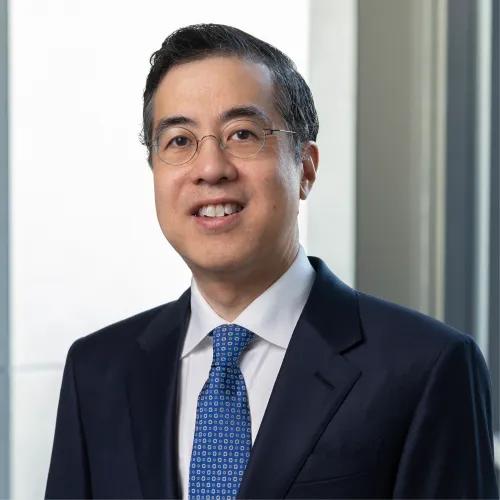 Professional headshot of a smiling man wearing round glasses, a navy suit, white shirt, and patterned blue tie, posed against a softly blurred office background.