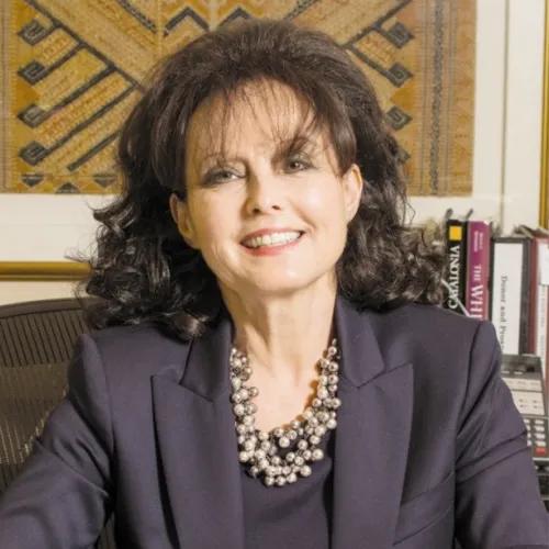  Professional portrait of a smiling woman with dark, shoulder-length hair, wearing a dark blazer and layered metallic necklace, seated in an office with framed textile art and books behind her.