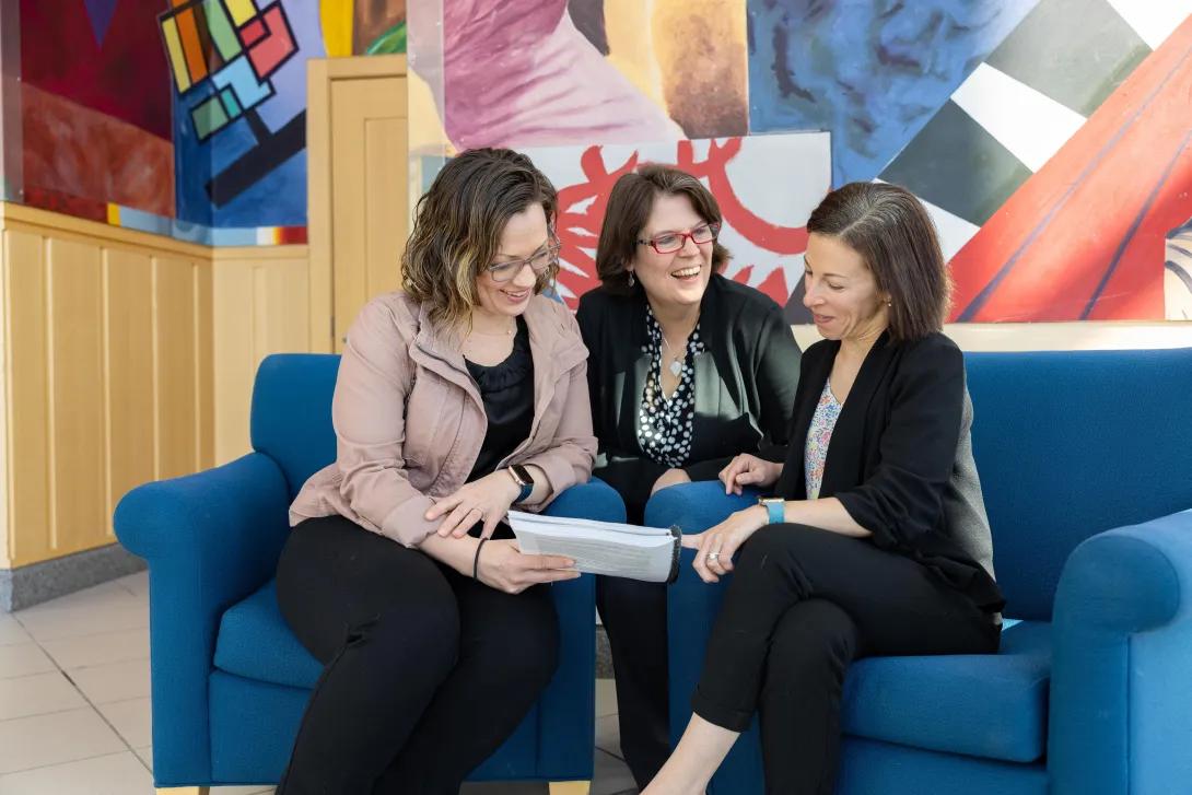  women sit on couch together and laugh while reading papers 