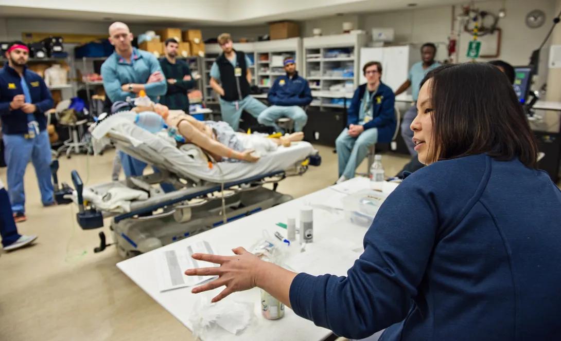 A medical simulation lab scene where a diverse group of people in scrubs attentively observe a woman explaining around a table, next to a patient mannequin on a bed.