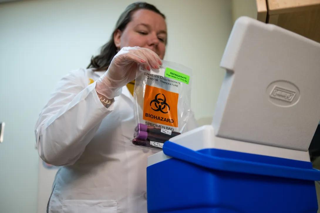 A lab technician in a white coat places a biohazard bag with blood samples into a blue cooler
