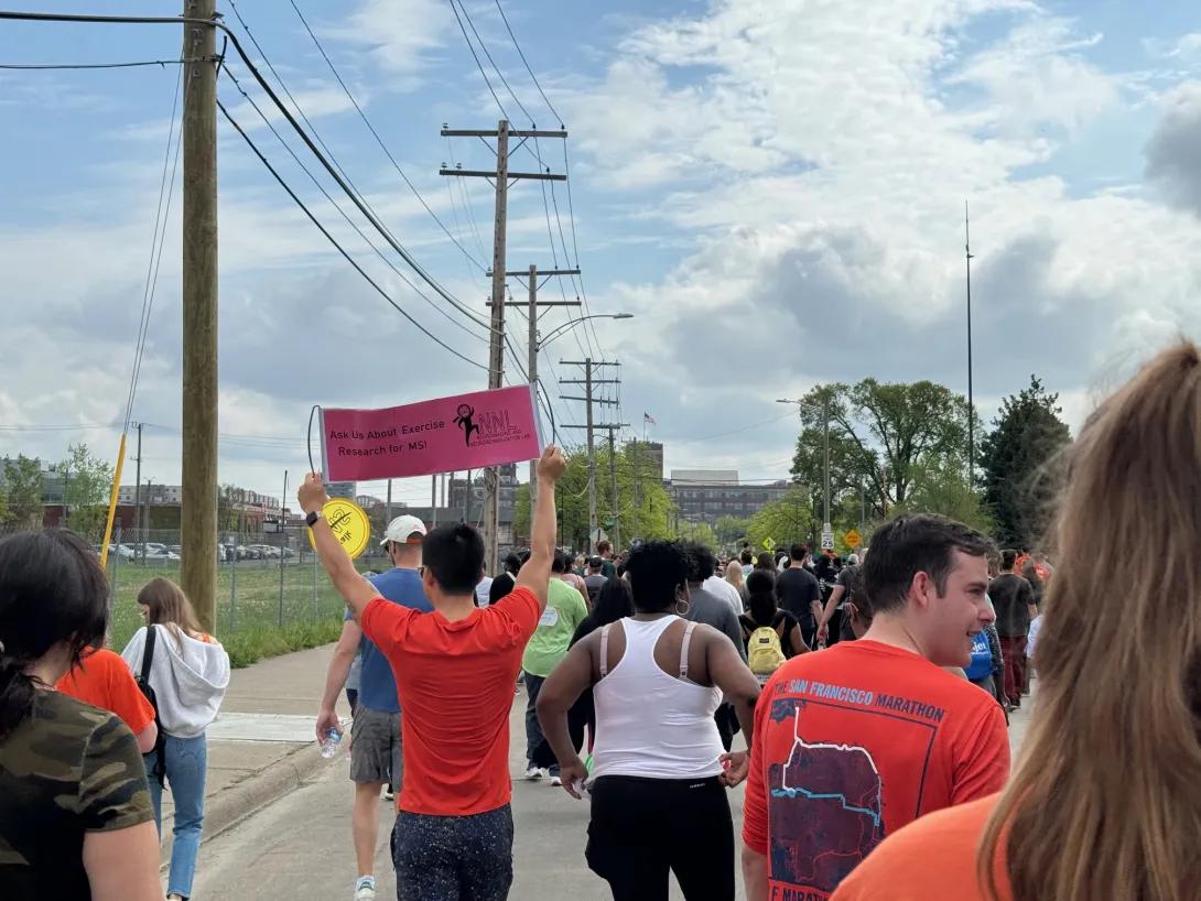 people walking during Walk MS, with one person holding up a sign above their head that says, "Ask Us About Exercise Research for MS!"