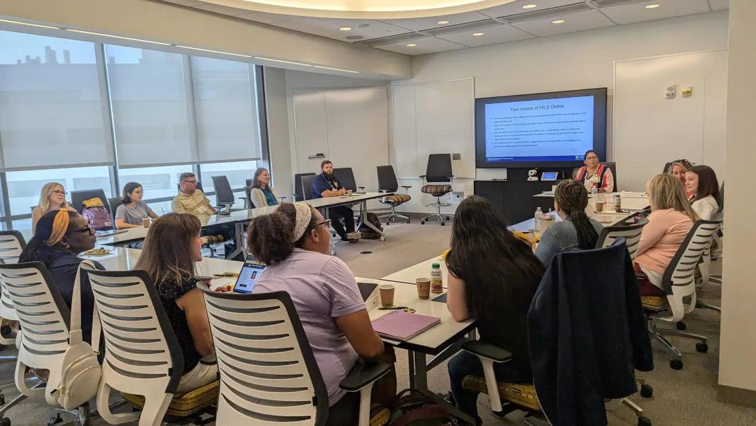 A group of participants seated around a conference table in a bright meeting room, attentively listening to a presentation displayed on a large screen at the front.