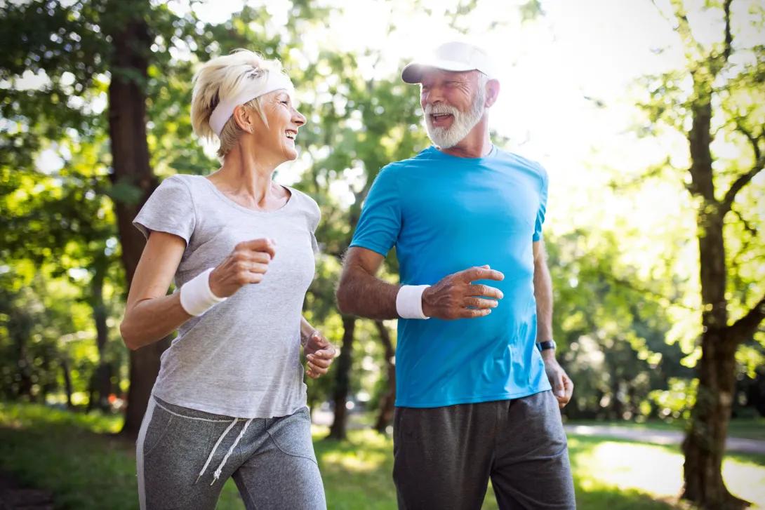 an older man and women briskly walking in a forest.  they are both dressed in workout wear
