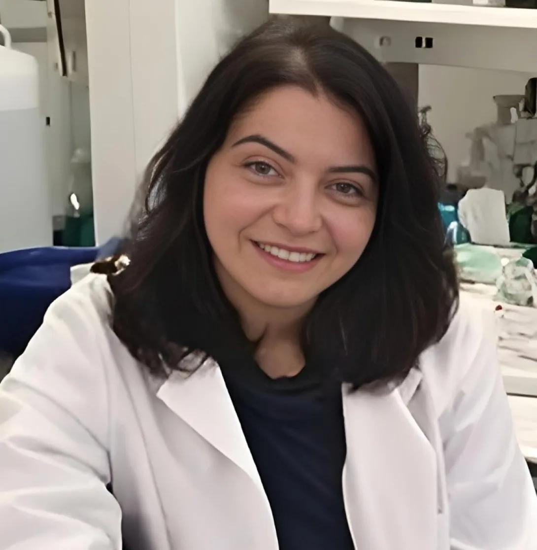 a Middle Eastern woman with shoulder length brown hair and a white coat 