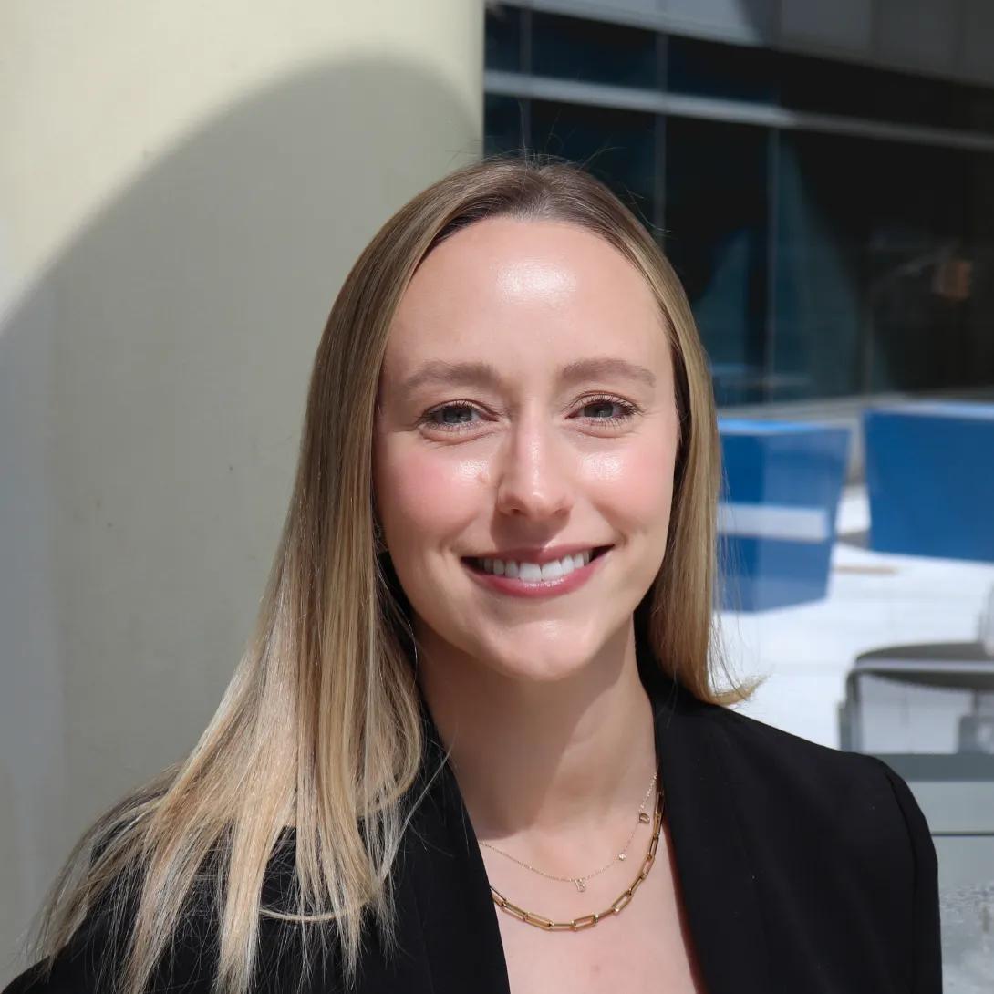 A person with long, straight blonde hair is wearing a black blazer and layered gold necklaces. The background shows an outdoor setting with a light-colored column and a modern building with blue panels and windows.