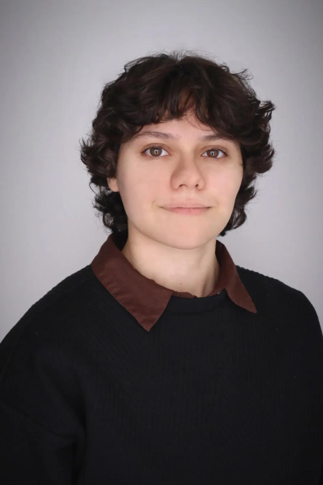 Person with short, wavy dark hair wearing a dark sweater over a brown collared shirt, positioned against a plain light gray background.