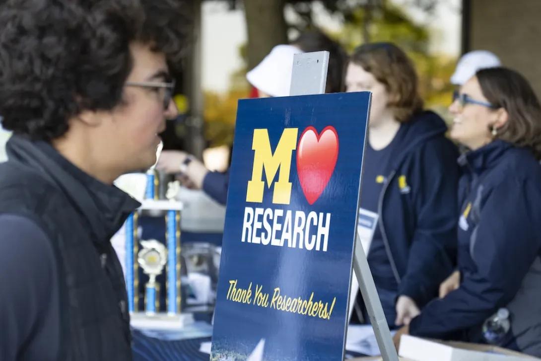 Photo og a person in front of the office of research tent with a sign being displayed M+Heart+Research