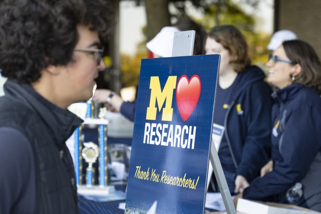 Photo og a person in front of the office of research tent with a sign being displayed M+Heart+Research