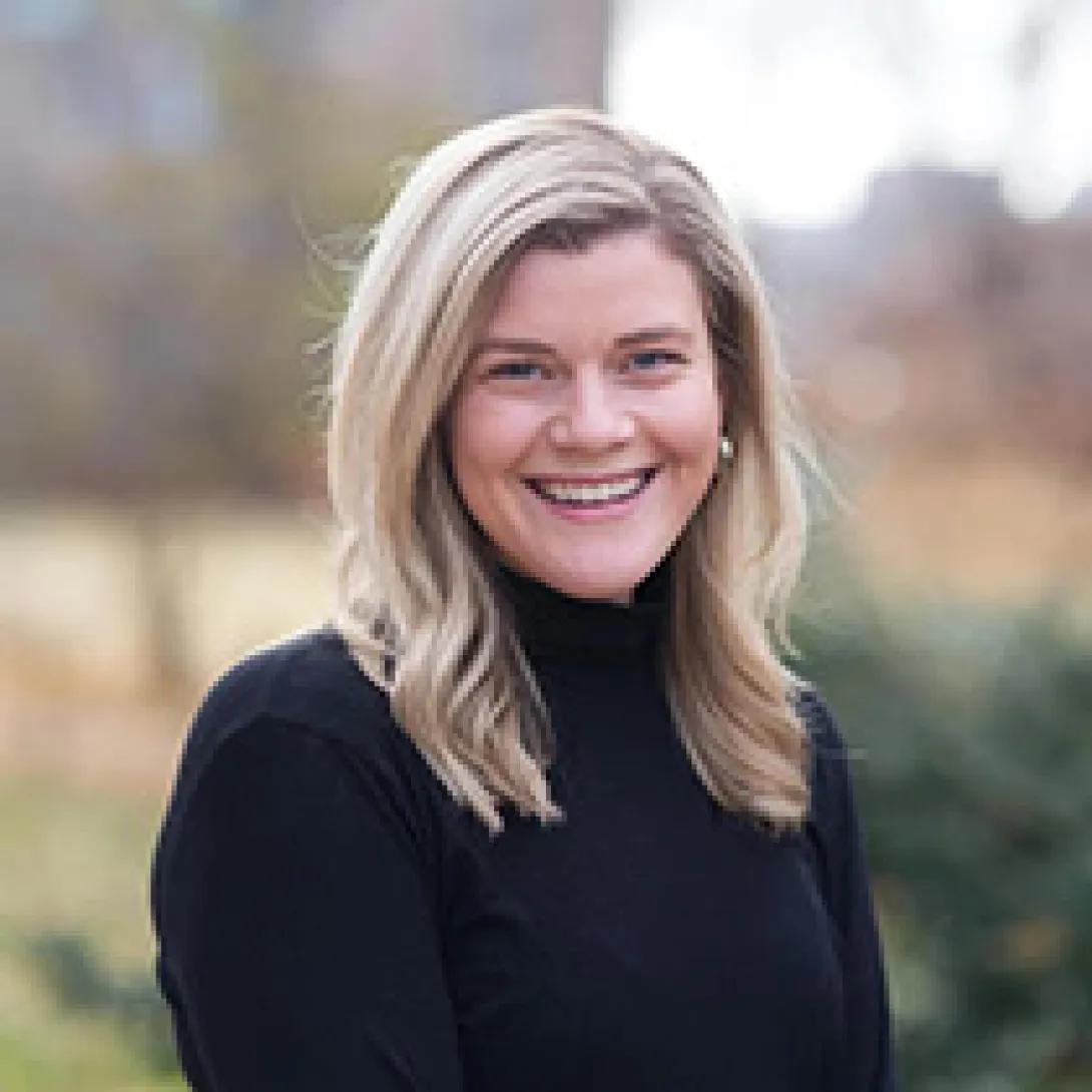 Headshot on woman outdoors, smiling with blonde hair in black turtleneck