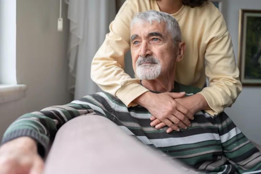 photo of a woman standing with her arms around a seated older man looking out into space