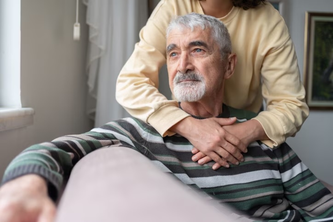 photo of a woman standing with her arms around a seated older man looking out into space