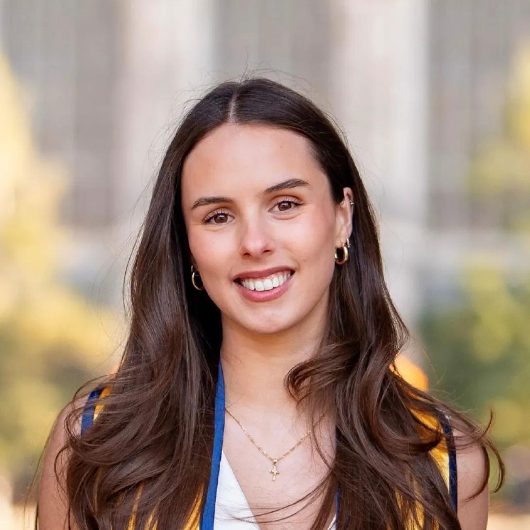 A person with long brown hair wearing a graduation stole with royal blue and gold trim, a white top, gold hoop earrings, and a gold necklace with a cross pendant, standing outdoors with a blurred background featuring greenery and a large building.