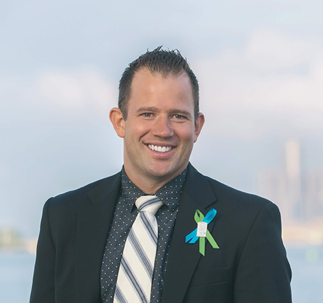 Smiling headshot of Luke Sparkman. He has blue and green awareness ribbons pinned to his lapel and is wearing a dark suit jacket.