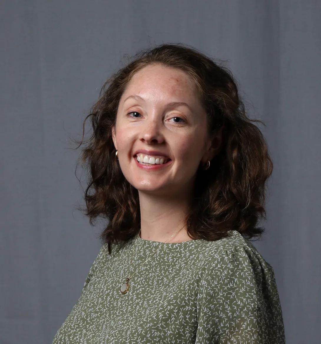 Headshot of person with shoulder-length brown hair, in green sweater, smiling in front of a gray background