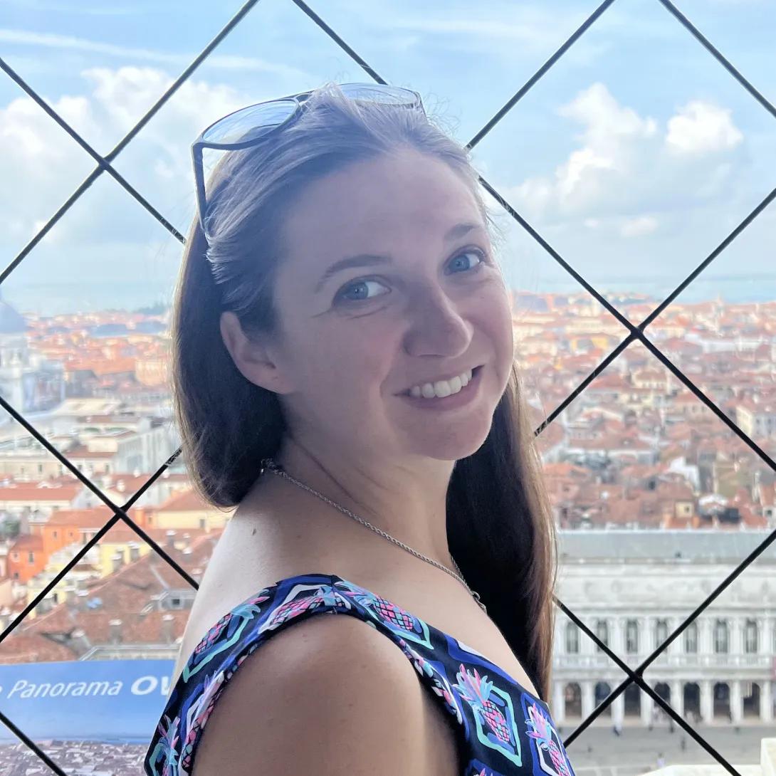Woman smiling, turned from the side, in front of a fenced look-out point with a city in the background