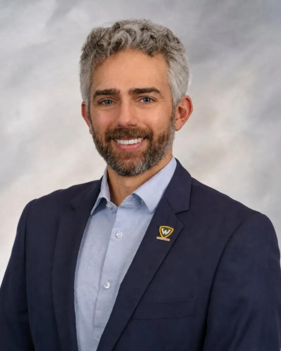 Professional headshot of a person smiling with gray hair and a beard, wearing a navy blazer with a small lapel pin and a light blue button-down shirt, posed against a gray studio background.