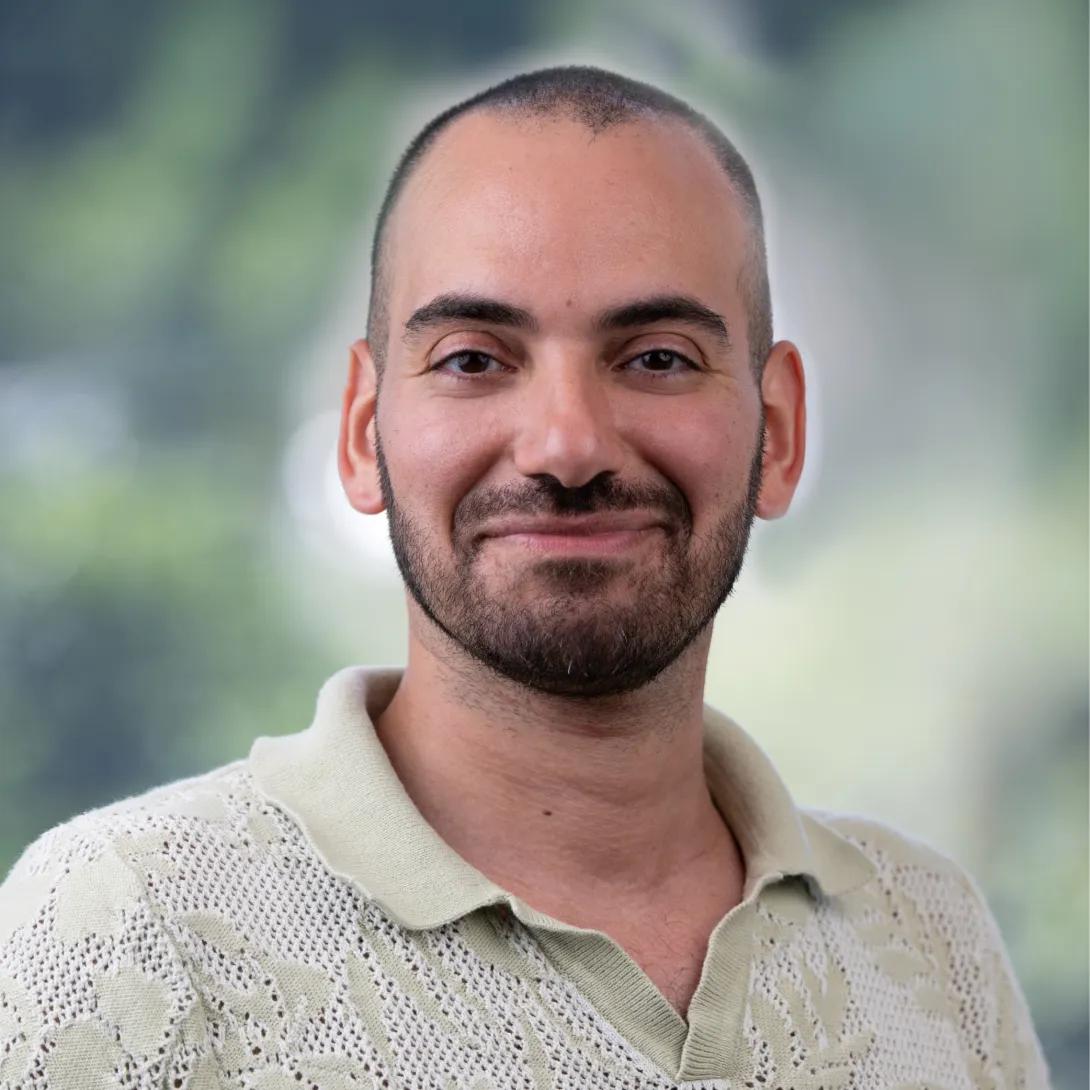 Head-and-shoulders portrait of a man with closely cropped hair and a short dark beard, smiling gently at the camera in a cream knit polo shirt against a softly blurred green background.