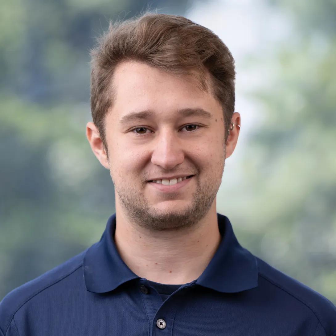 Head-and-shoulders portrait of a young man with short brown hair and light facial hair, smiling at the camera in a navy polo shirt against a softly blurred green background.