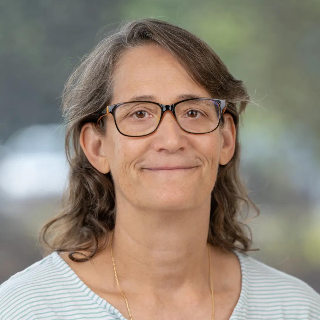 Professional headshot of a woman with shoulder-length brown hair and dark-framed glasses, smiling against a softly blurred background.