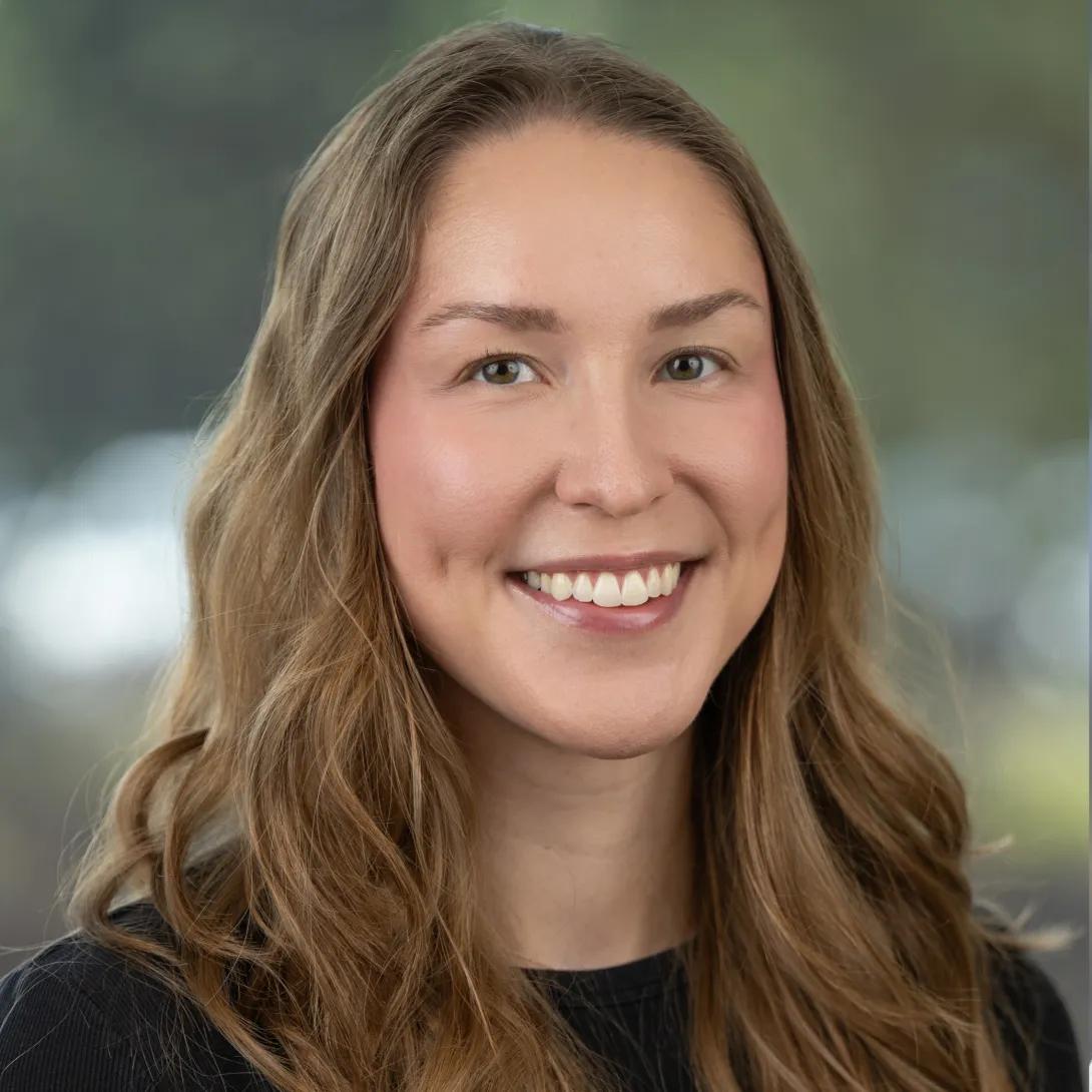Professional headshot of a woman with long, wavy light-brown hair wearing a black top and smiling against a softly blurred background.