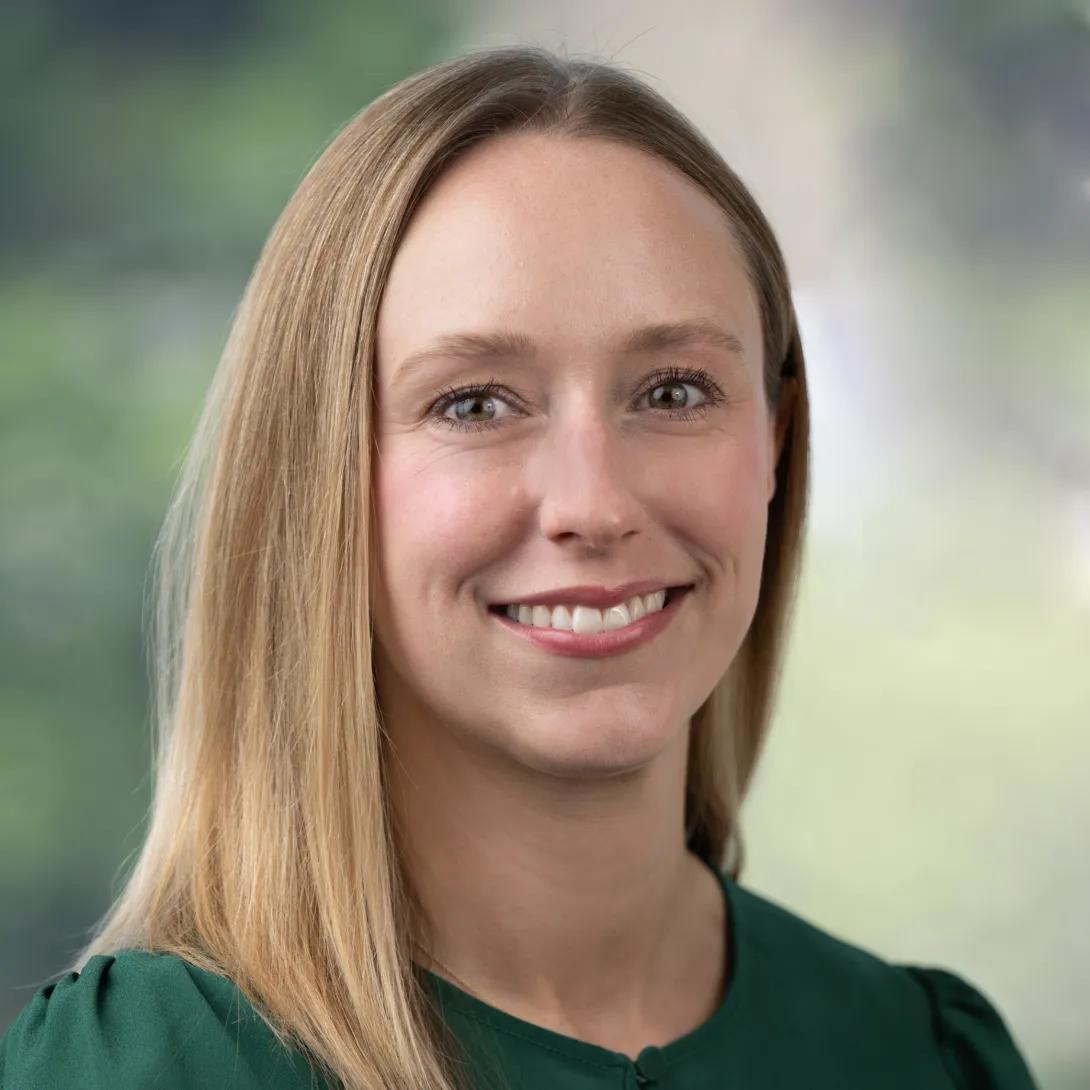 Head-and-shoulders portrait of a woman with long, straight blonde hair, smiling at the camera in a dark green top against a softly blurred green background.