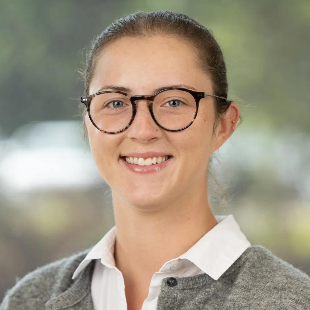 Professional headshot of a smiling woman with her brown hair pulled back, wearing round tortoiseshell glasses, a white collared shirt, and a gray cardigan against a softly blurred background.