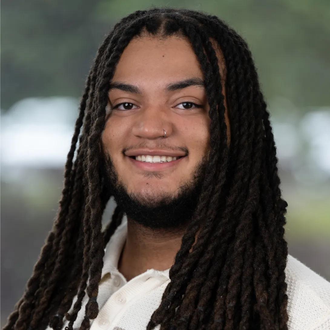 Professional headshot of a smiling man with long dark locs, a short beard, and a silver nose ring, wearing a cream-colored shirt against a softly blurred background.