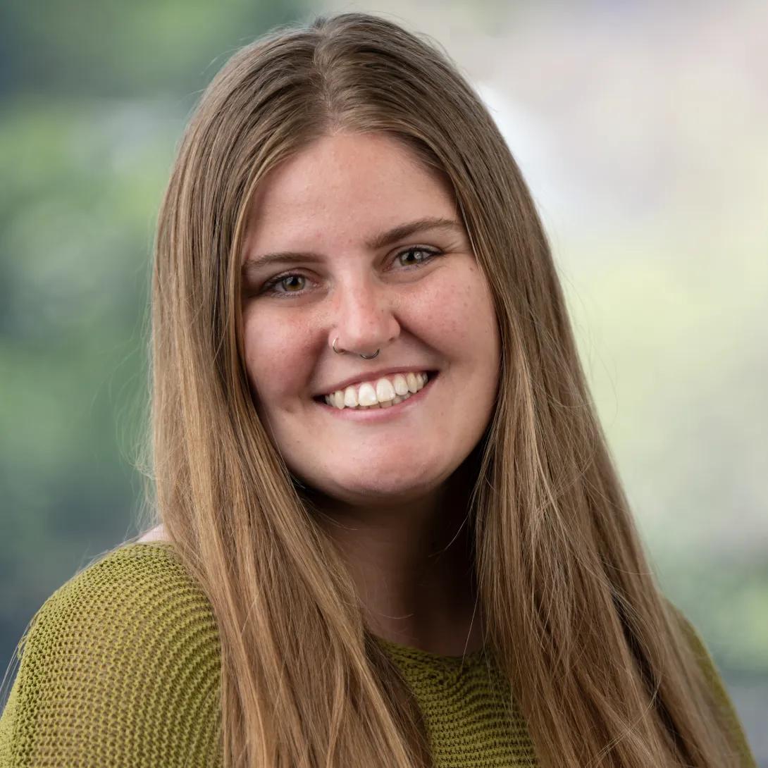 Professional headshot of a smiling person with long, straight light-brown hair, two silver nose rings, and a green knit top against a softly blurred background.