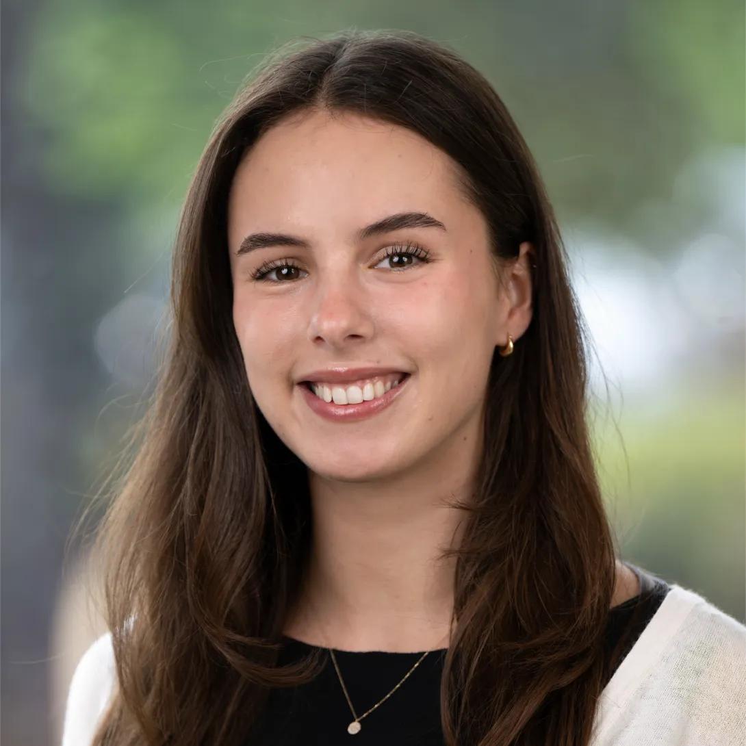 Professional headshot of a smiling woman with long, straight brown hair, wearing gold hoop earrings, a pendant necklace, and a cream cardigan over a black top against a softly blurred background.