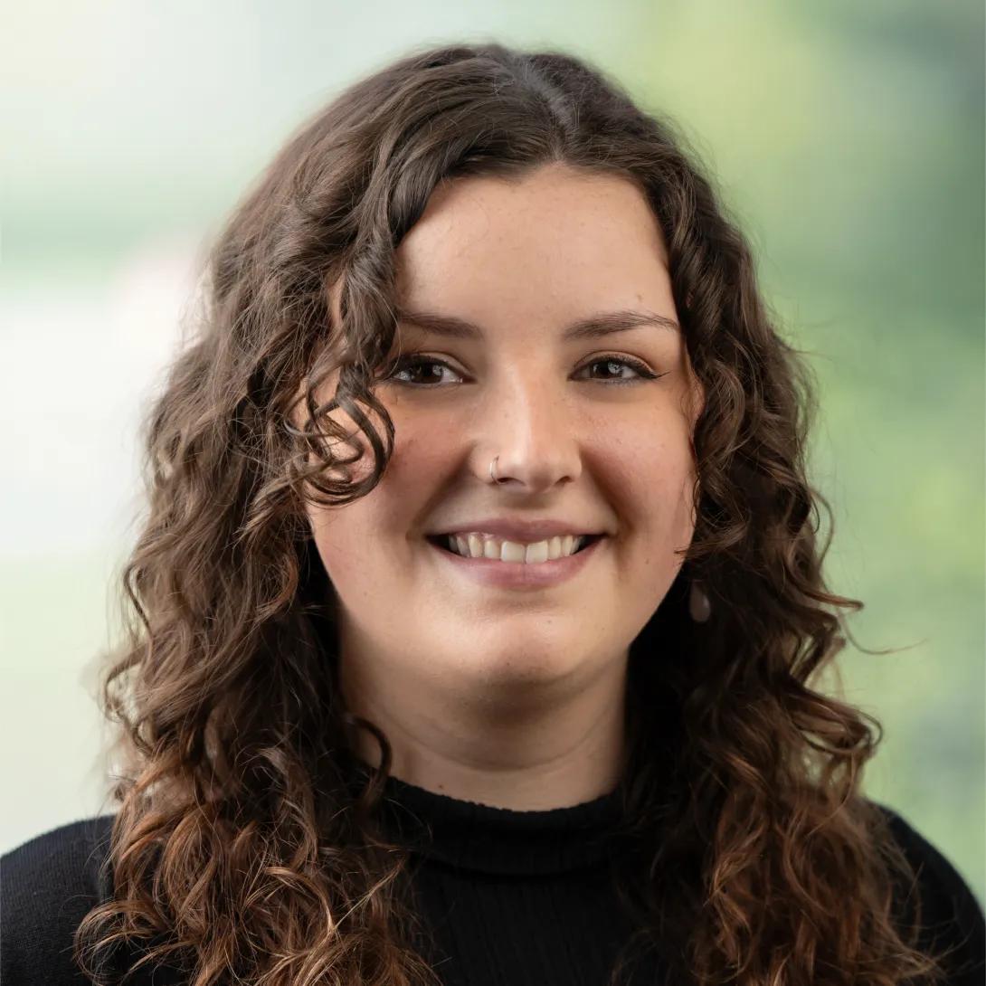 Head-and-shoulders portrait of a smiling woman with long, curly brown hair, a nose ring, and drop earrings, wearing a black top against a softly blurred green background.