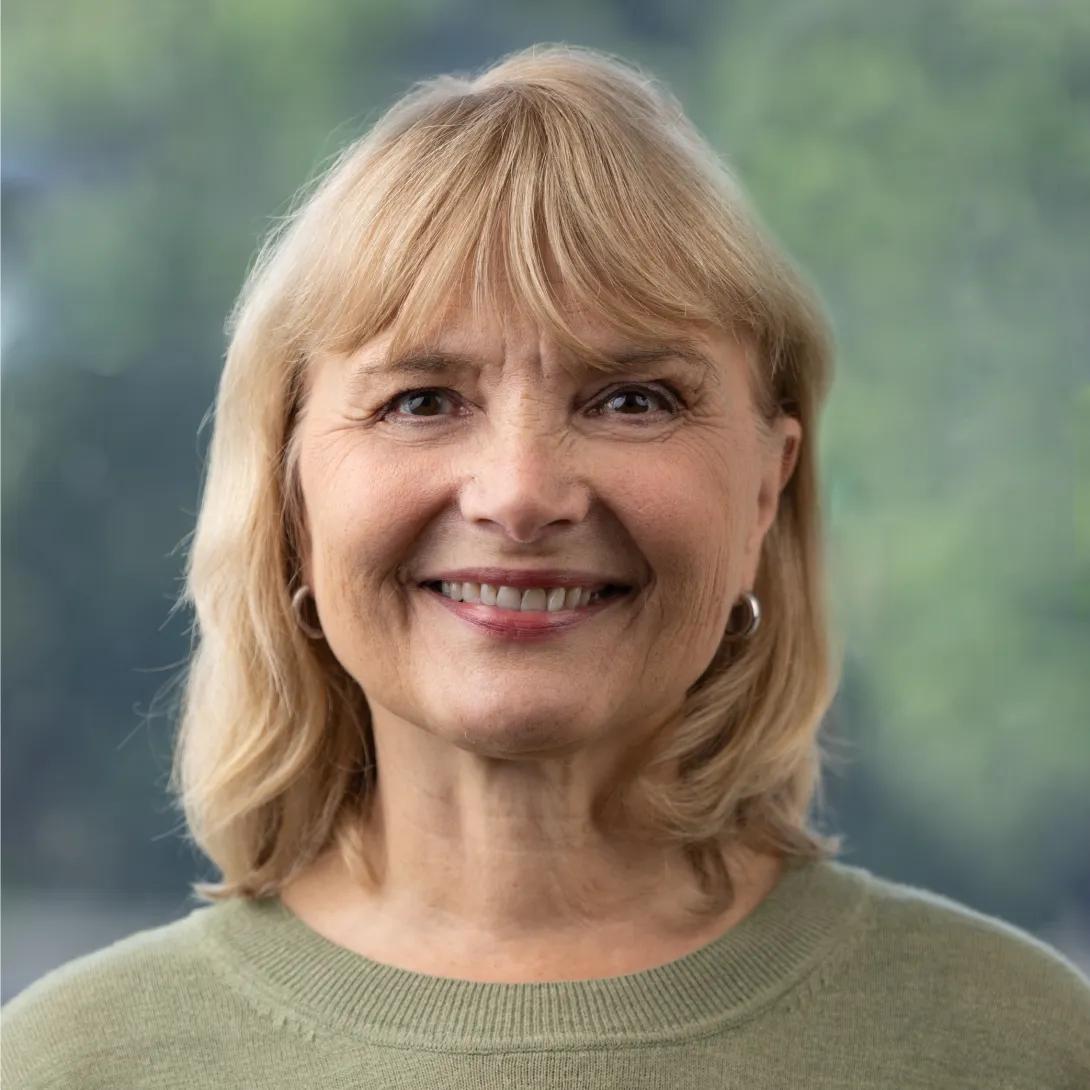 Head-and-shoulders portrait of a woman with shoulder-length blonde hair and bangs, smiling at the camera in a sage-green sweater against a softly blurred green background.